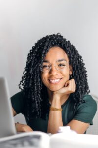 Joyful businesswoman with curly hair smiling at camera while using laptop indoors.